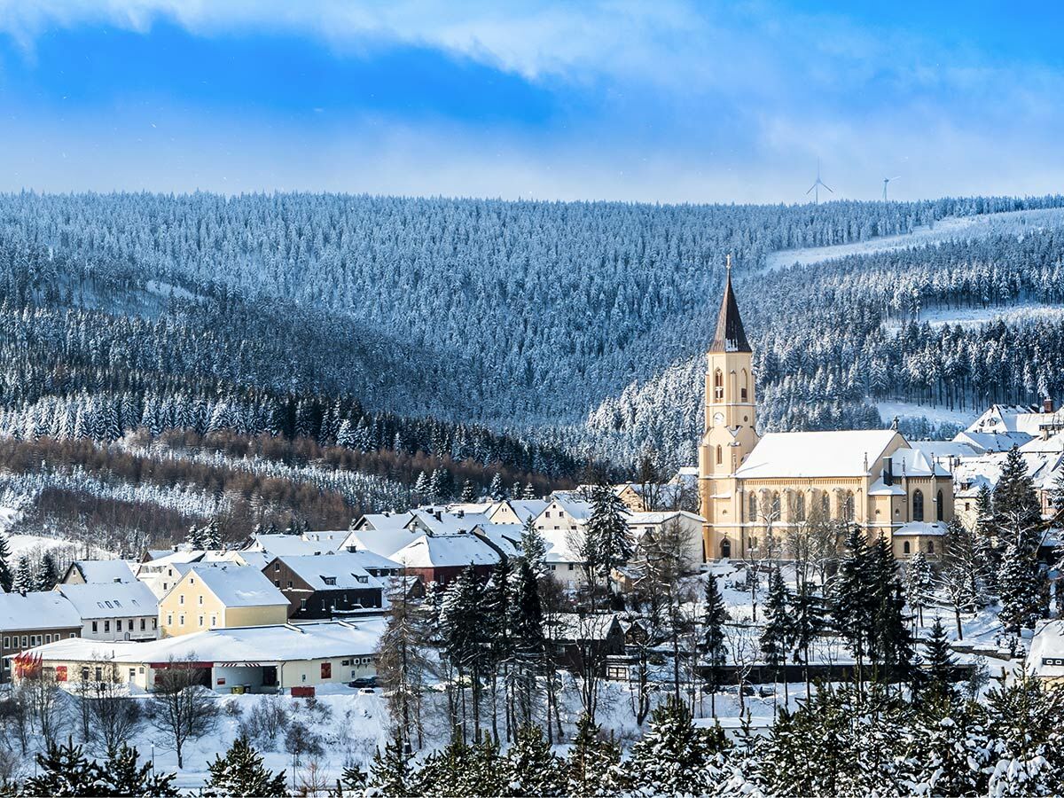 Verschneites Dorf mit markanter Kirche und Häusern, eingebettet in tief verschneite Tannenwälder unter blauem Himmel.