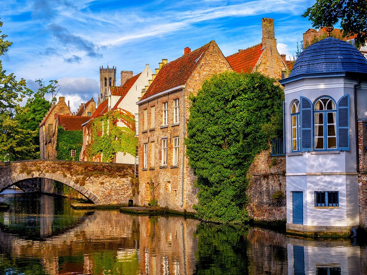 Malerischer Kanal in Brügge, Belgien, mit historischen Häusern, Steinbrücke und markantem weißen Gebäude mit blauer Kuppel.