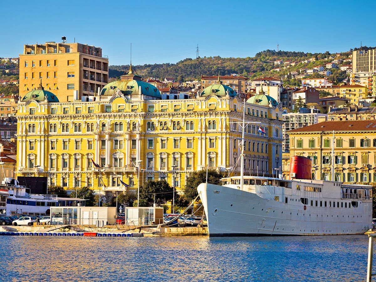 Blick auf die historische Küstenstadt Opatija, Kroatien, mit prächtigem gelbem Hotel, Schiff am Hafen und blauem Meer.