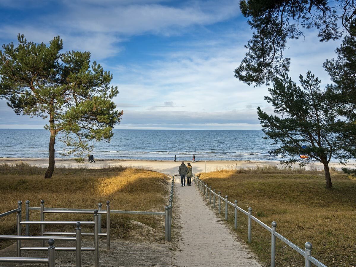Paar geht auf Sandweg durch Dünen zum herbstlichen Ostseestrand mit blauem Meer und Himmel. Kiefern stehen am Wegesrand.
