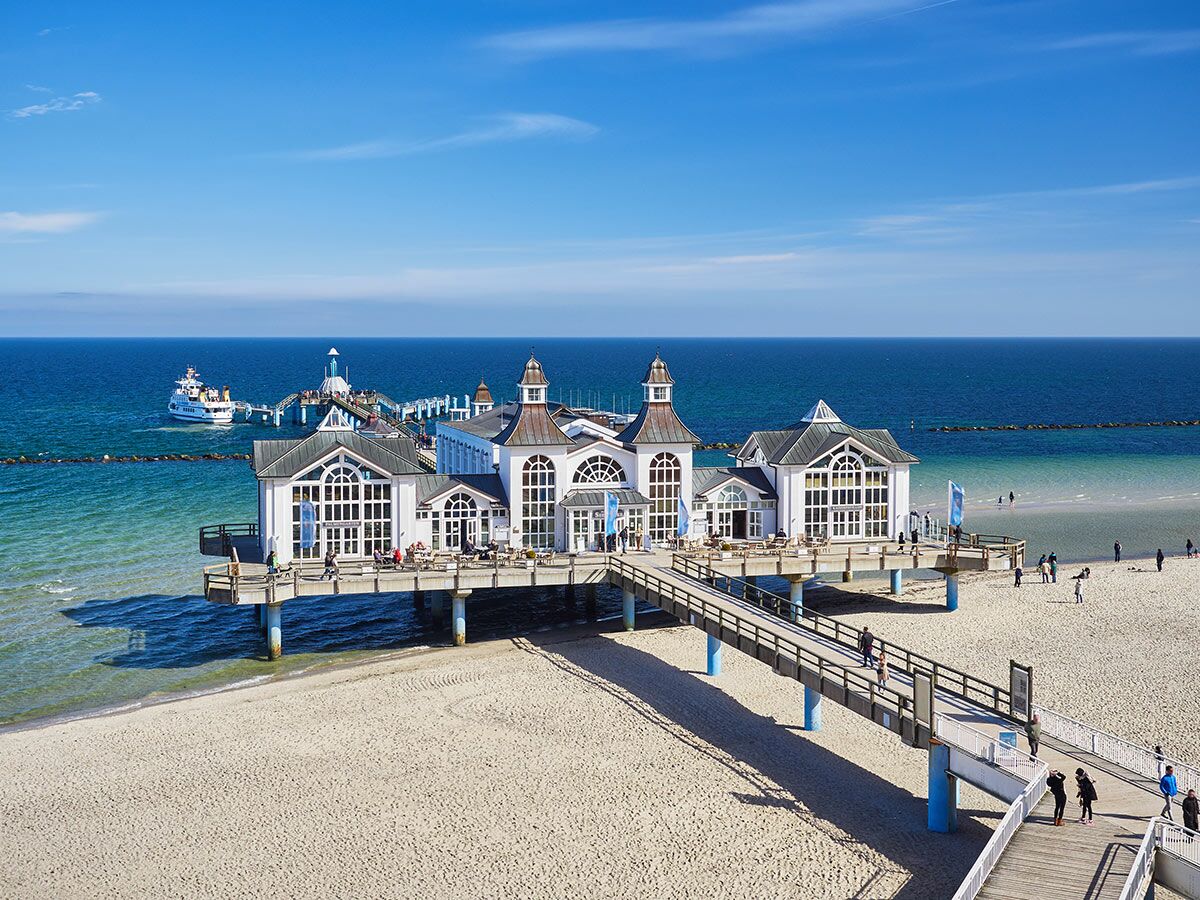 Panoramablick auf die historische Seebrücke Sellin an der Ostsee mit ihren markanten Gebäuden, einem Ausflugsschiff und sonnigem Sandstrand.