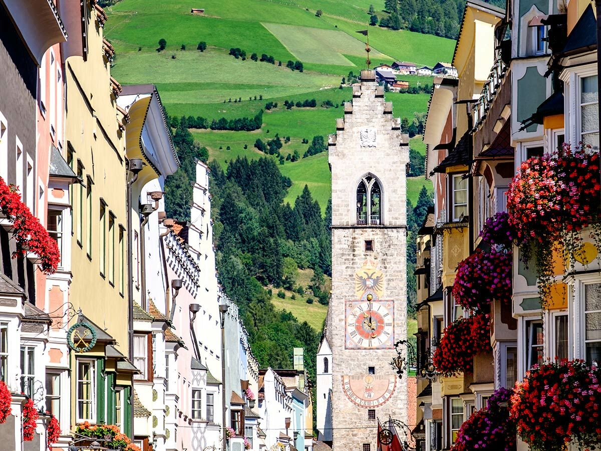 Malerische Alpenstadt-Straße mit farbenfrohen Häusern, roten Blumenkästen, historischem Glockenturm und grünen Berghängen.