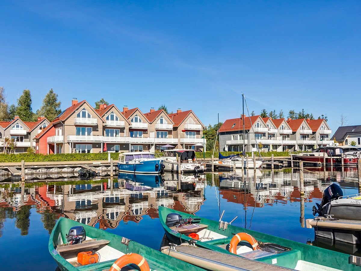 Blick auf einen Kanal mit bunten Ferienhäusern und vielen Booten im Hafen, idyllisch im klaren Wasser gespiegelt unter blauem Himmel.