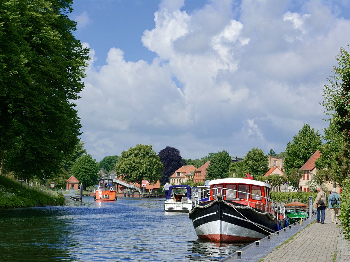 Malerische Kanalansicht mit bunten Booten, grünen Bäumen und Häusern unter blauem Himmel. Zwei Personen spazieren am Uferweg.