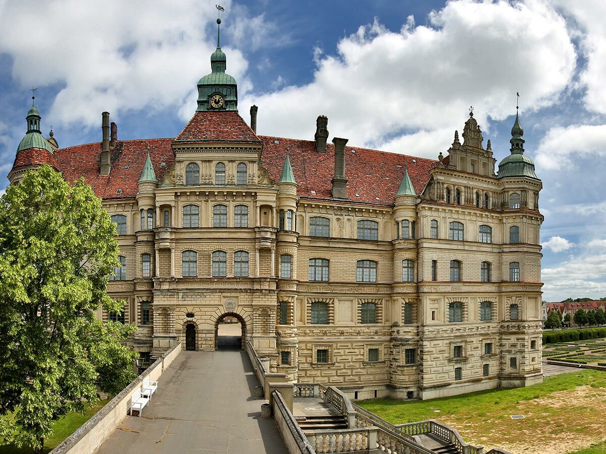 Blick auf das imposante Schloss Güstrow mit roten Dächern, grünen Türmen und einem Uhrenturm unter blauem Himmel.