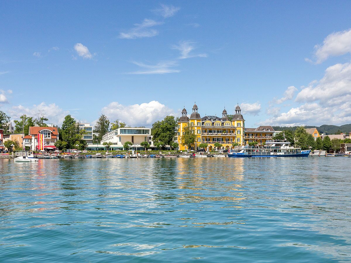 Panoramablick auf die Seepromenade Pörtschach am Wörthersee mit gelbem Schlosshotel, Booten und blauem Himmel.