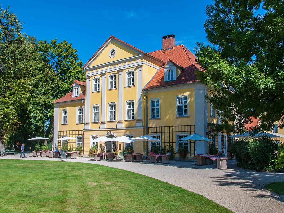 Gelbes Herrenhaus mit rotem Dach und Gartenterrasse. Gäste entspannen unter Sonnenschirmen auf grünem Rasen bei sonnigem Wetter.