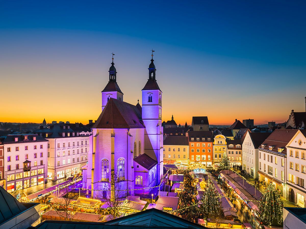 Weihnachtsmarkt in Regensburg bei Abenddämmerung mit lila beleuchteter Kirche und festlichen Ständen, umgeben von historischen Gebäuden.