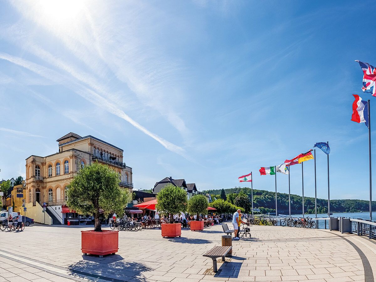 Panoramablick auf eine belebte Promenade mit vielen Flaggen, Cafés und Menschen an einem sonnigen Seeufer.