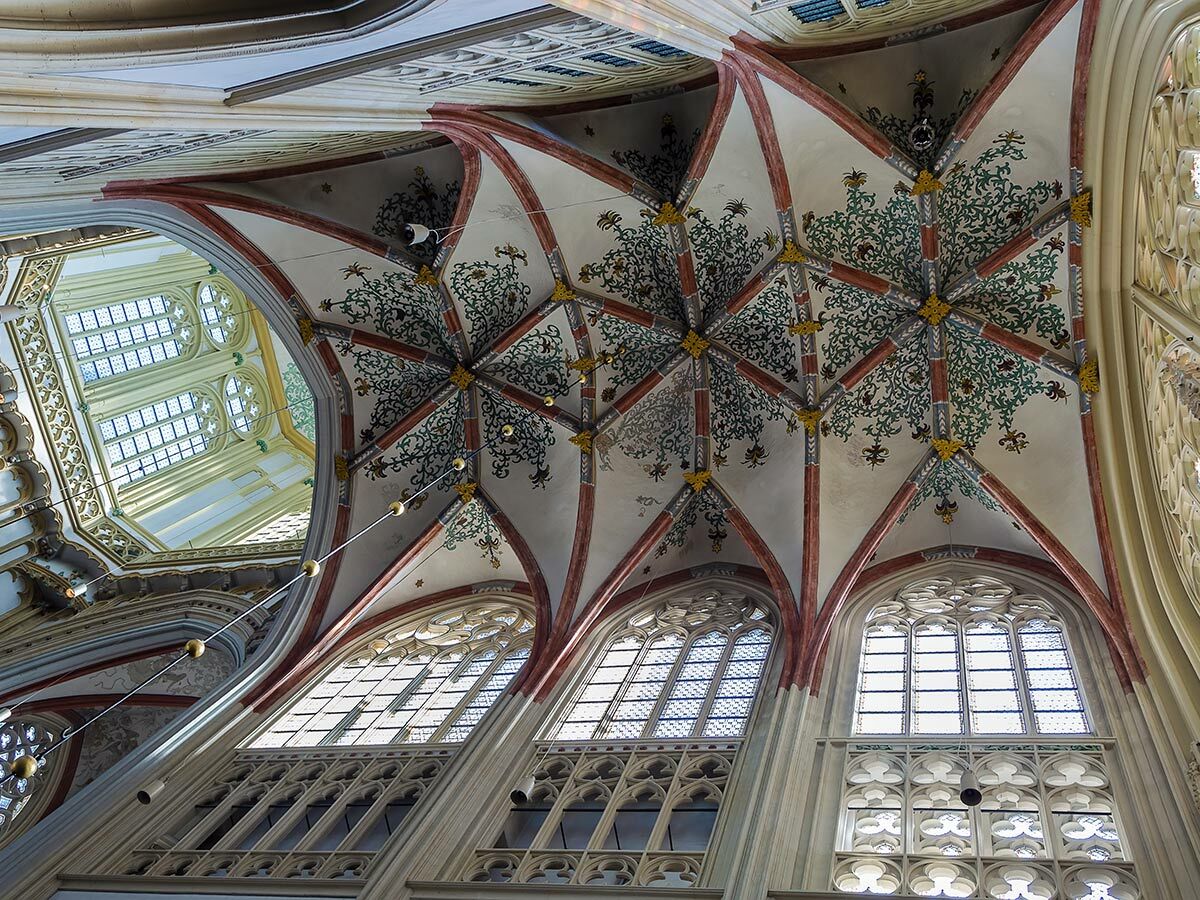 Blick nach oben in ein prächtiges gotisches Kirchengewölbe mit detaillierten Malereien, Rippen und hohen Fenstern.