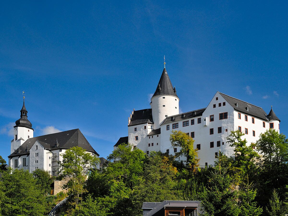 Historisches weißes Schloss Augustusburg mit dunklen Dächern und Türmen auf bewaldetem Hügel unter blauem Himmel. Sachsens Wahrzeichen.
