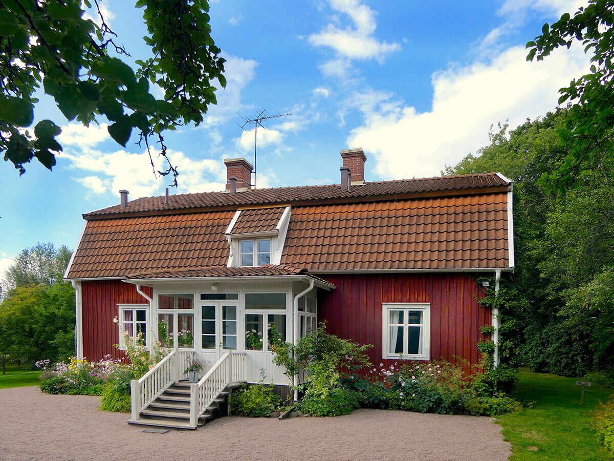 Idyllisches rotes Holzhaus mit weißer Veranda, Blumen und grünen Bäumen unter blauem Himmel.
