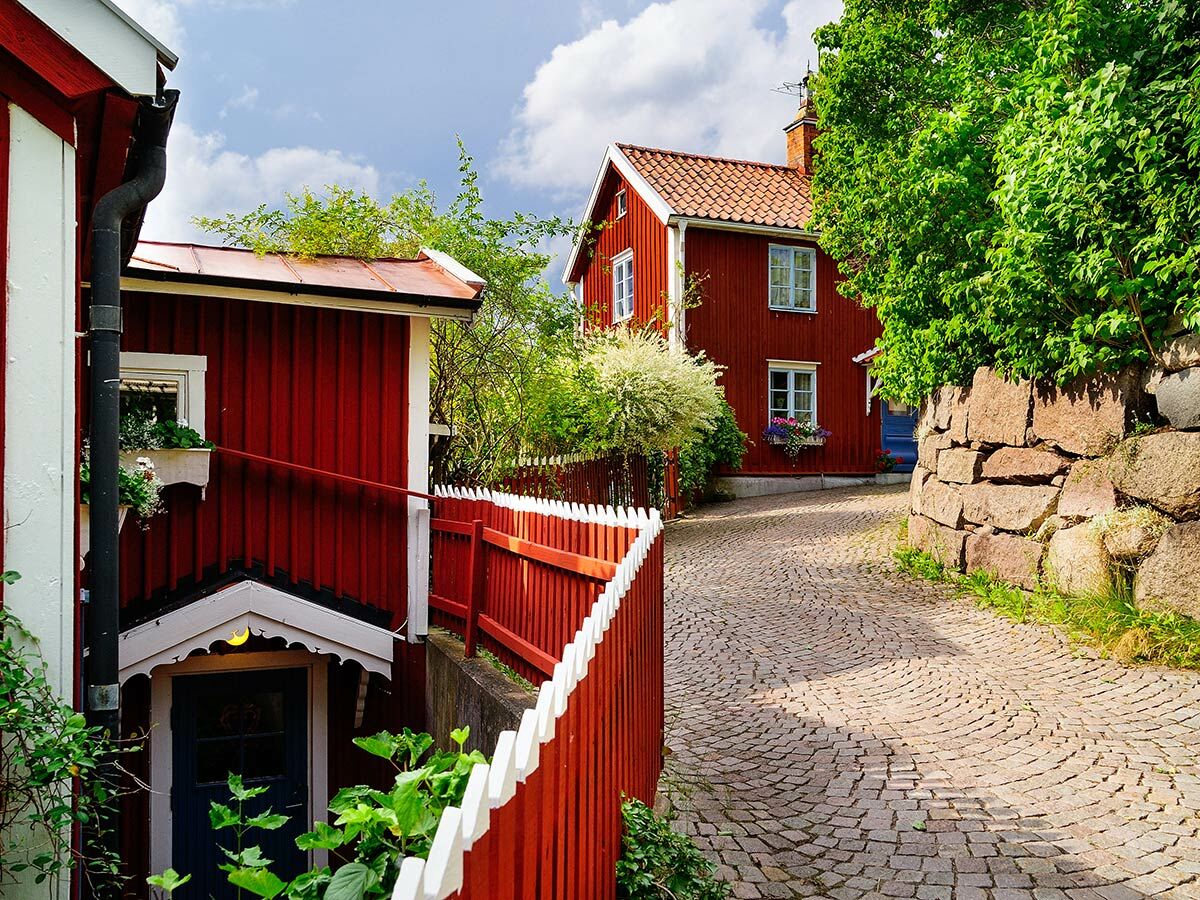 Charmante rote Holzhäuser mit weißem Zaun und gepflasterter Gasse, umgeben von Grün und einer Steinmauer.