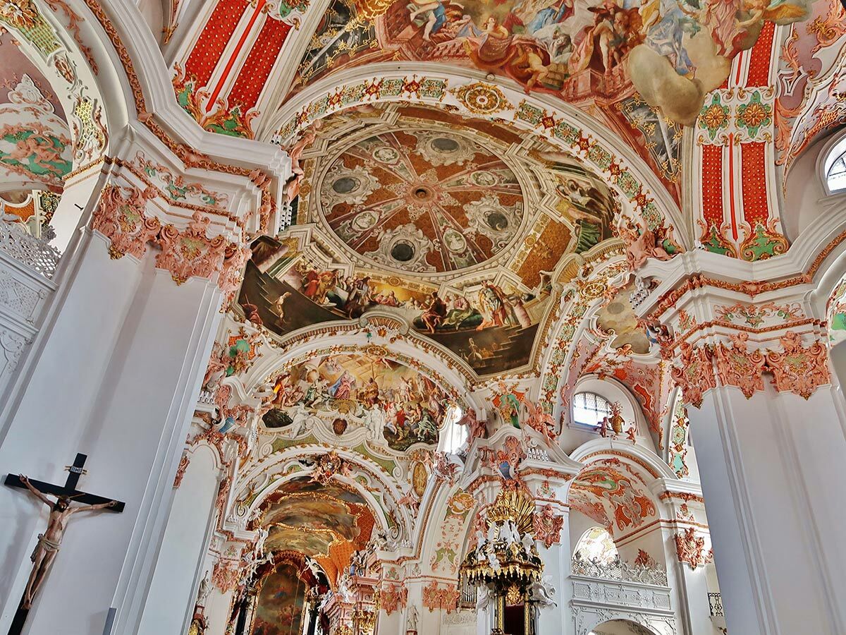 Prachtvoller barocker Kircheninnenraum mit detailreichen Deckenfresken, goldenem Stuck, rosa Verzierungen und weißen Säulen.