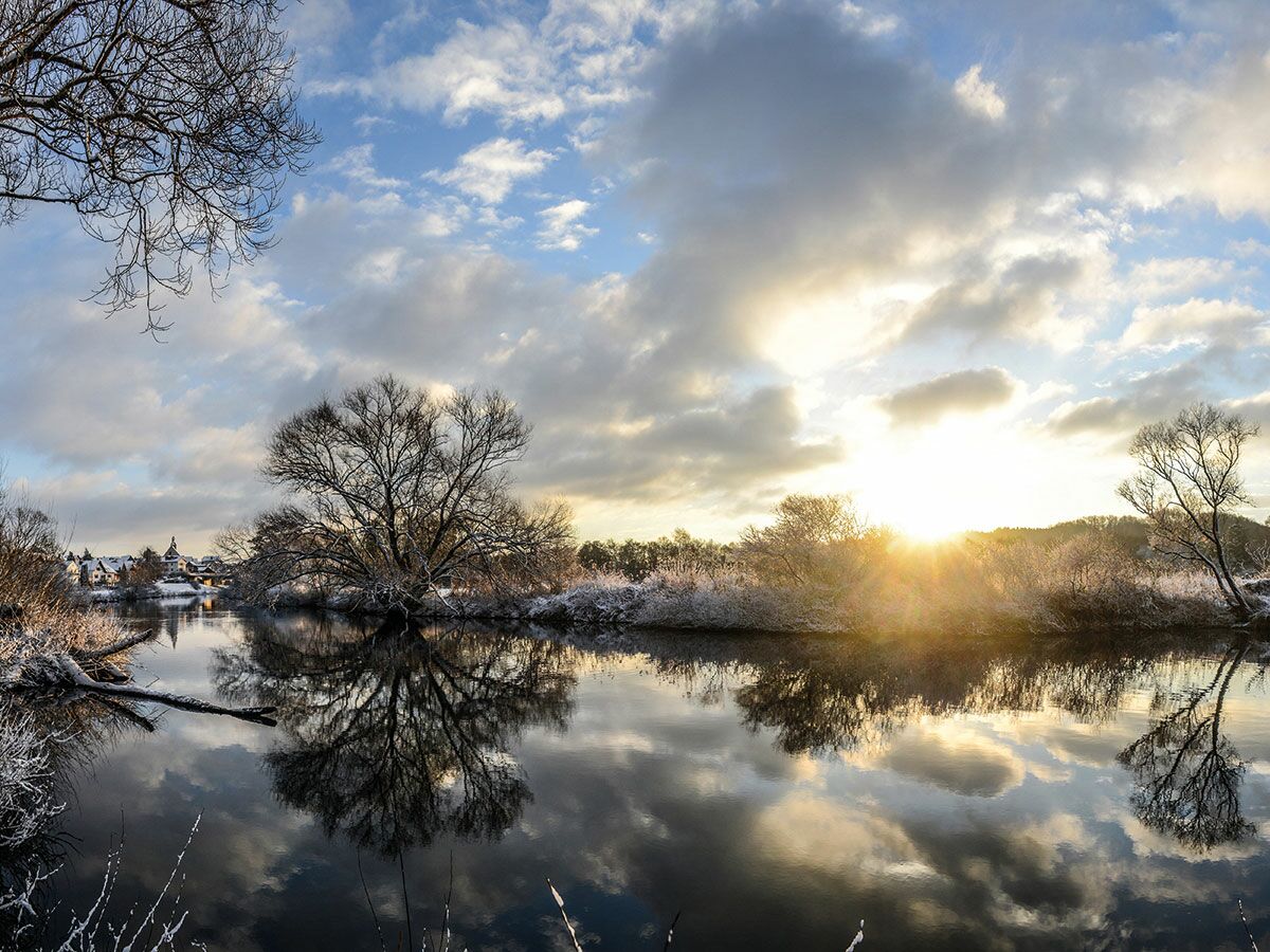 Idyllische Winterlandschaft mit reifbedeckten Bäumen an einem Fluss und einem strahlenden Sonnenaufgang, der sich im Wasser spiegelt.