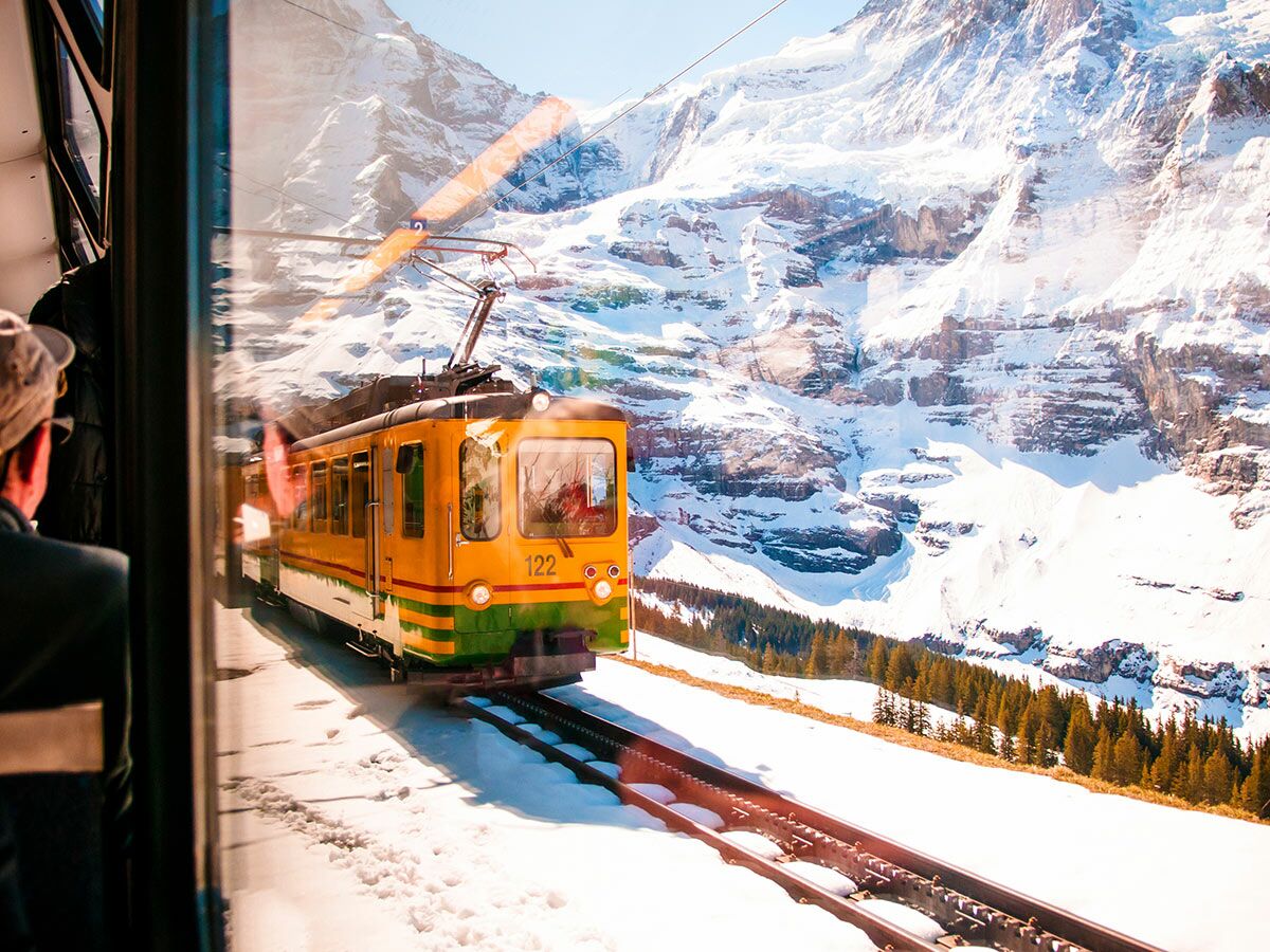 Gelbe Bergbahn hält an schneebedeckter Station vor majestätischen Alpen. Ansicht aus Fenster mit Reflexionen und Reisendem.