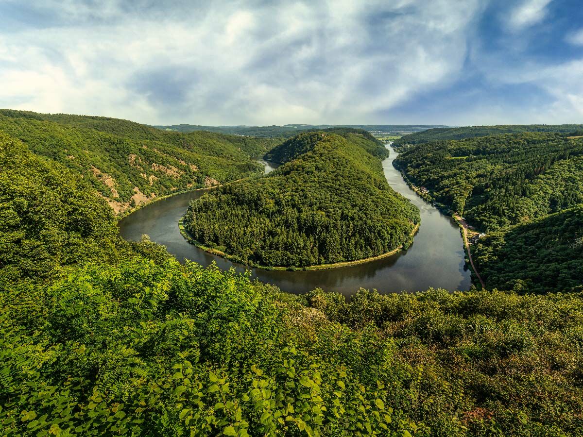 Luftaufnahme der Saarschleife, einem berühmten Flussmäander umgeben von dichten grünen Wäldern und Hügeln unter blauem Himmel.