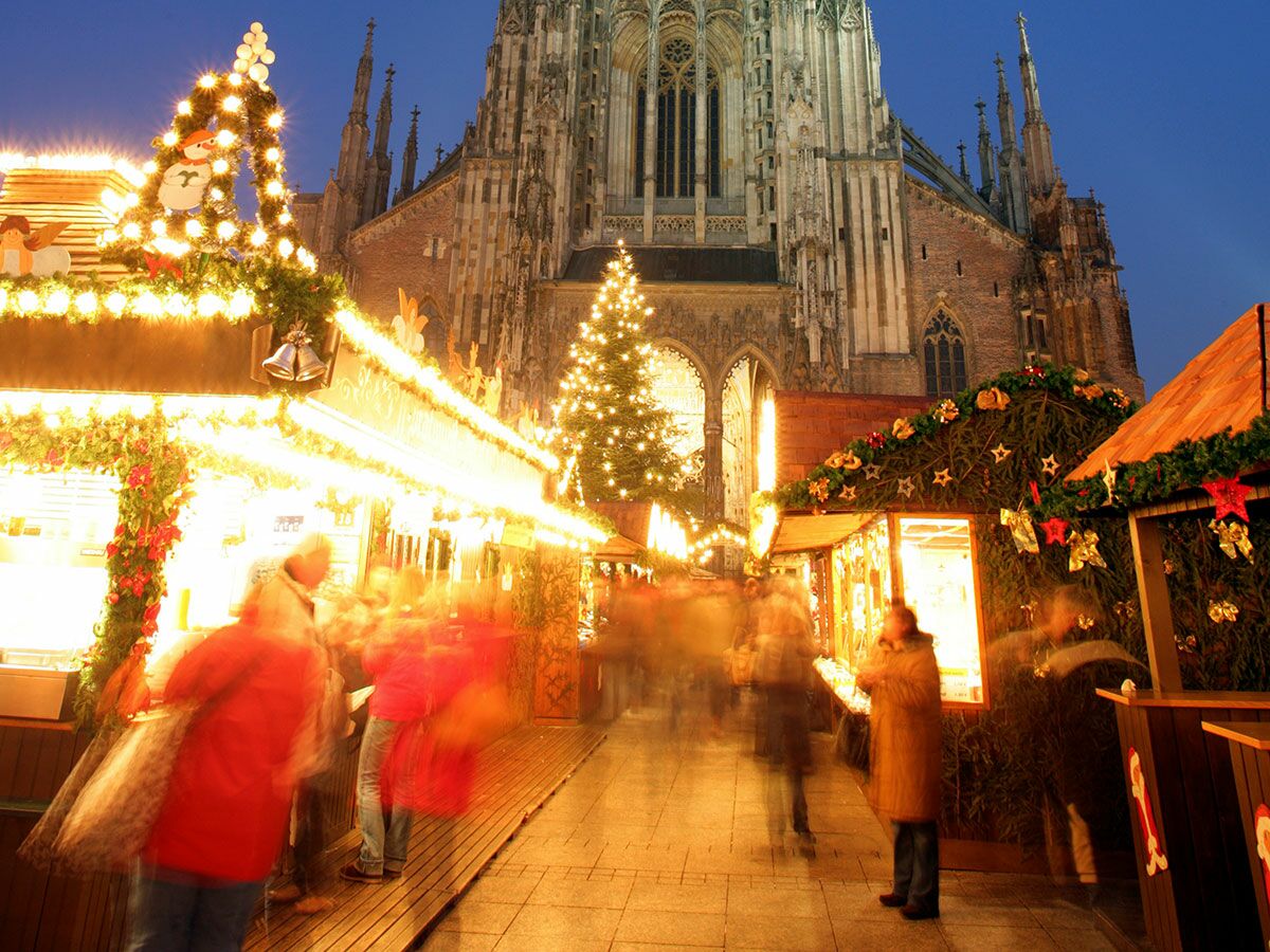 Festlicher Weihnachtsmarkt mit beleuchteten Buden, Weihnachtsbaum und verschwommenen Menschen vor dem Ulmer Münster bei Abenddämmerung.