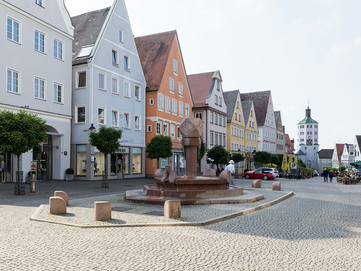 Bunter, belebter Stadtplatz mit historischen Giebelhäusern, einem Brunnen und einem Turm im Hintergrund, gepflasterter Boden.