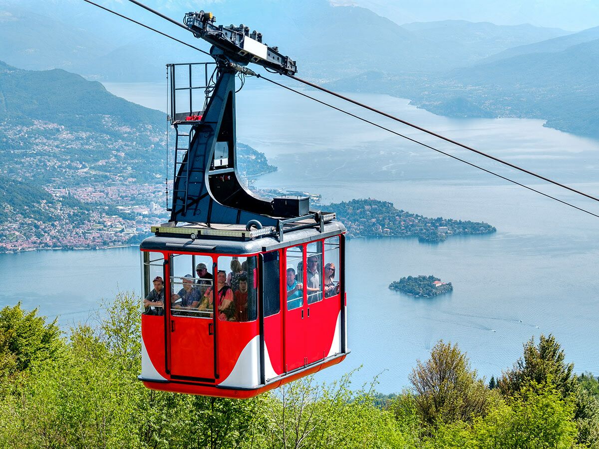 Rote Seilbahn voller Passagiere schwebt über einem großen, blauen See mit Inseln, umgeben von Bergen und grünen Bäumen.