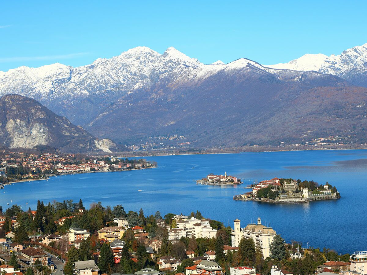 Panoramablick auf den Lago Maggiore mit den Borromäischen Inseln, Gebäuden und Gärten, umgeben von schneebedeckten Alpen und Uferstädten.