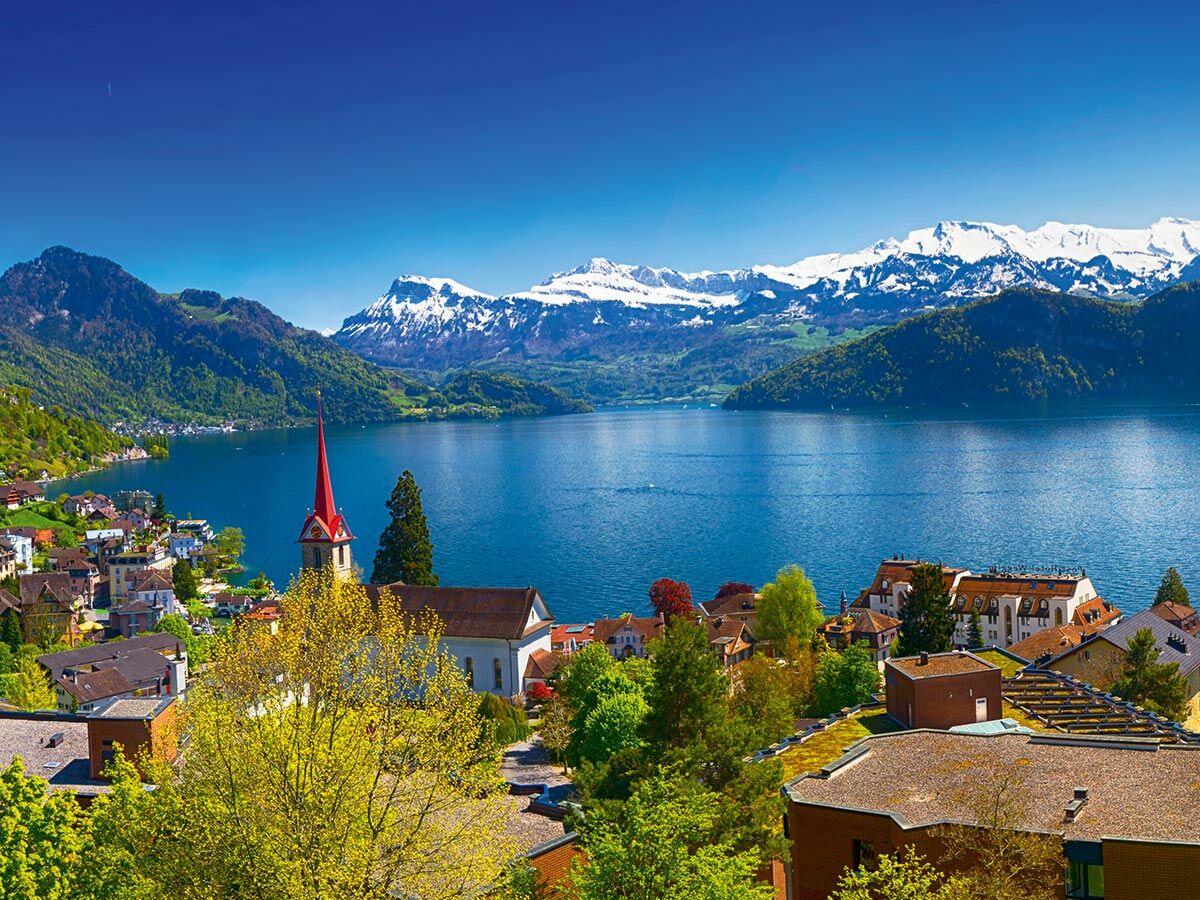 Panoramablick auf idyllisches Schweizer Dorf am See, umgeben von grünen Bergen und schneebedeckten Alpen, mit markanter Kirche.