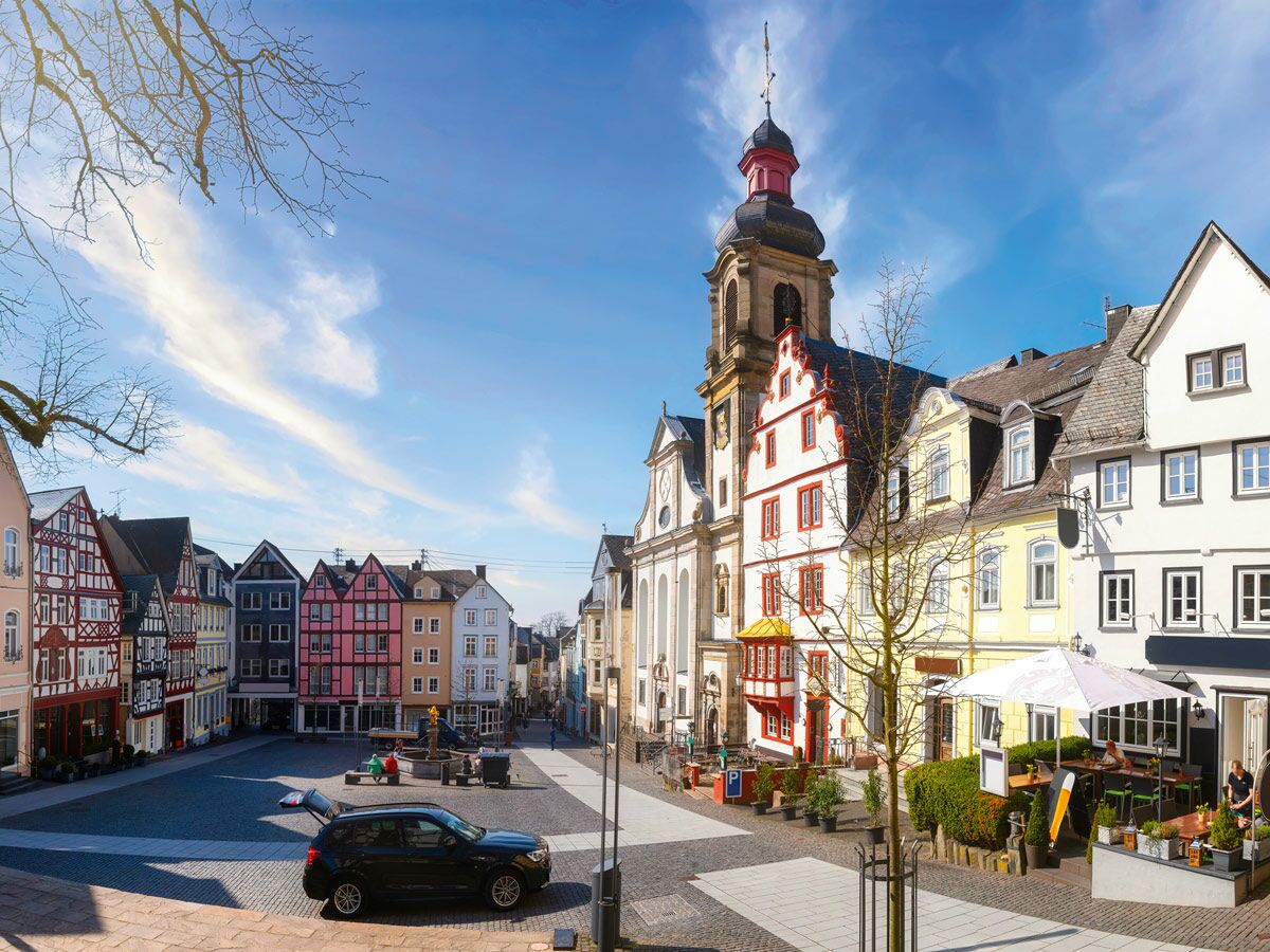 Sonniger historischer Stadtplatz mit farbenfrohen Fachwerkhäusern, markanter Kirche und belebter Außengastronomie.