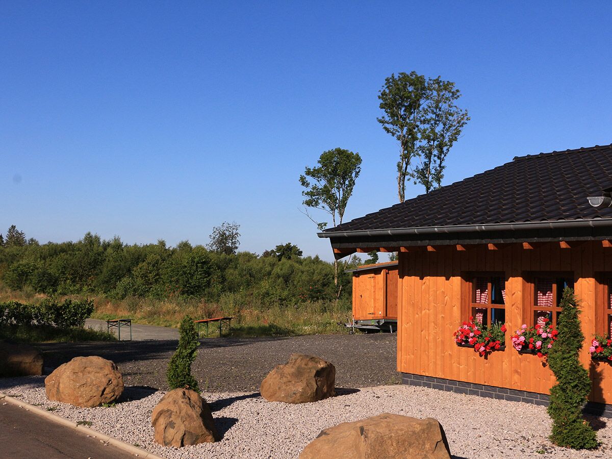 Gemütliches Holzhaus mit roten Blumenkästen, umgeben von Kies, Felsen und grüner Natur unter blauem Himmel.