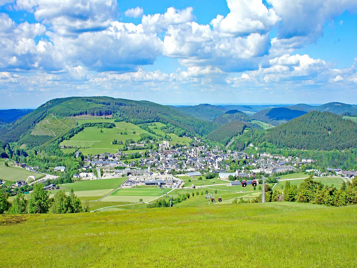 Panoramablick auf ein von grünen Wäldern und Wiesen umgebenes Tal mit einer Stadt, Seilbahn und blauem Himmel.