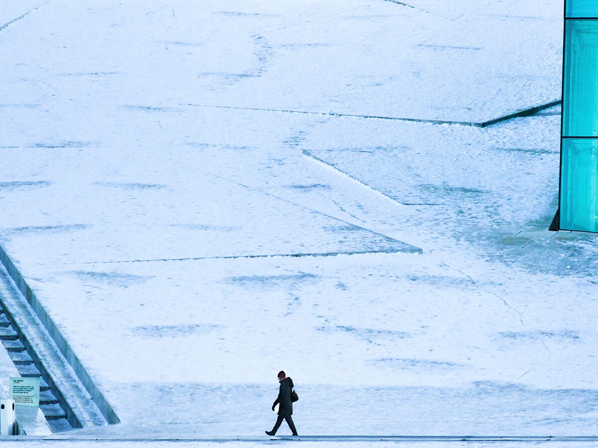 Eine einsame Person geht durch eine weite, eisige Winterlandschaft mit blauen Akzenten und einer Treppe im Vordergrund.