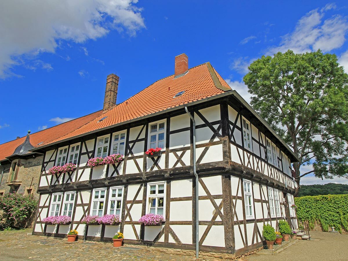 Traditionelles Fachwerkhaus mit rot gedecktem Dach, weißen Fenstern und rosa Blumenkästen unter blauem Himmel.