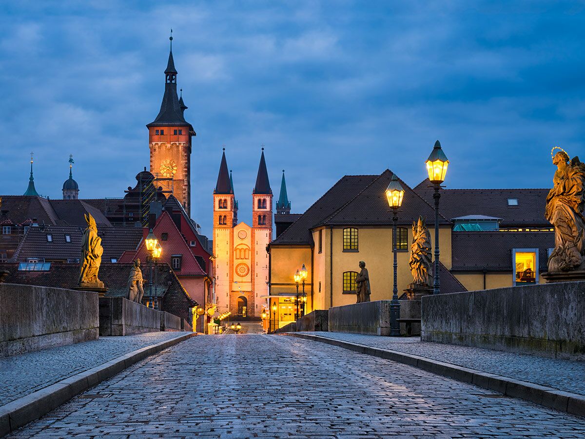 Abendliche Ansicht der Alten Mainbrücke in Würzburg mit beleuchteten Statuen, Kopfsteinpflaster, Altstadtgebäuden und Kirchtürmen bei Dämmerung.