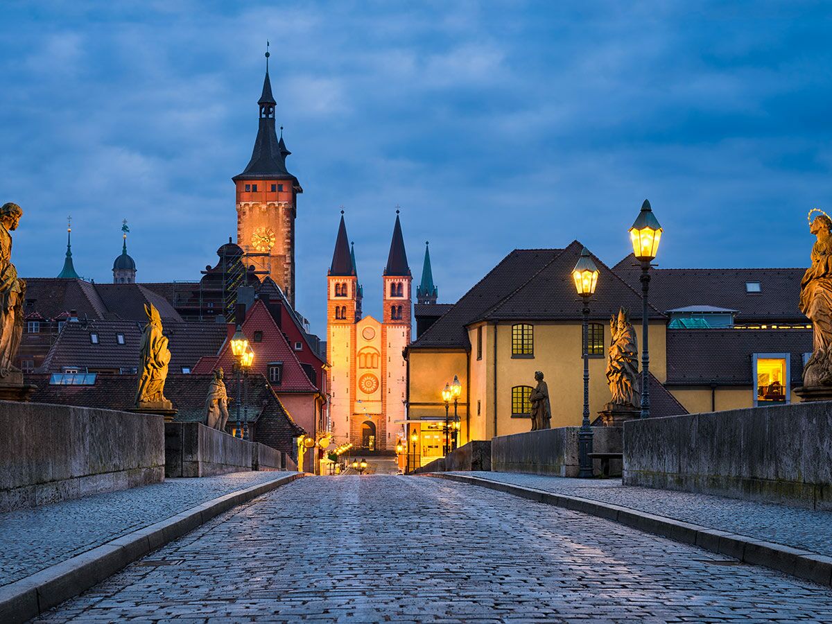 Abendansicht der Alten Mainbrücke in Würzburg mit Barockstatuen, beleuchteten Gebäuden und Kirchtürmen unter blauem Himmel.