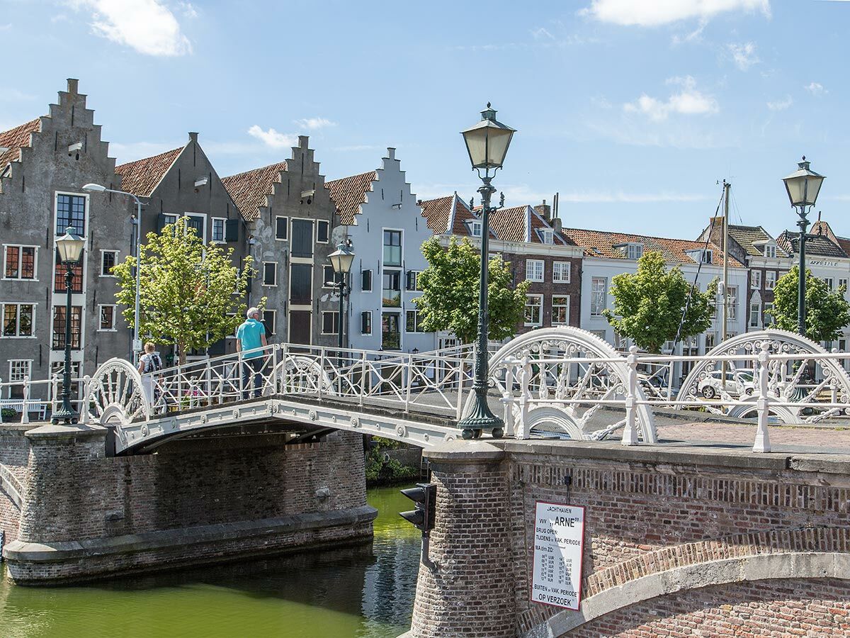 Weiße, verzierte Grachtenbrücke über Kanal in historischer niederländischer Stadt mit Giebelhäusern und Spaziergängern am sonnigen Tag.