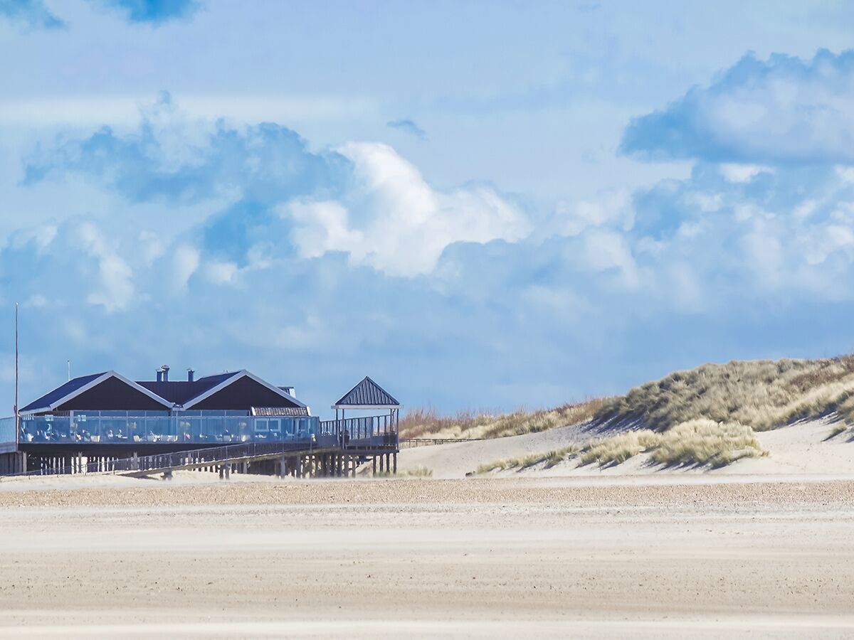 Modernes Restaurant auf Holzstelzen am weiten Sandstrand mit Dünen und blauem Himmel.