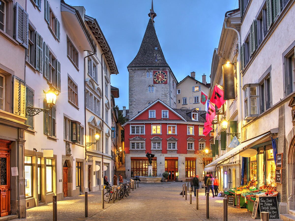 Historische Zürcher Altstadt-Gasse am Abend mit beleuchteten Gebäuden, Pflastersteinen, Passanten und dem Peterskirche-Turm.