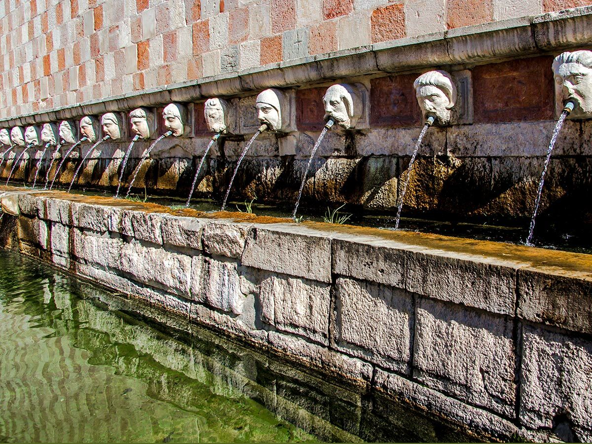 Historischer Steinbrunnen mit einer Reihe von Skulpturen menschlicher Köpfe, aus deren Mündern Wasser in ein langes Becken fließt.