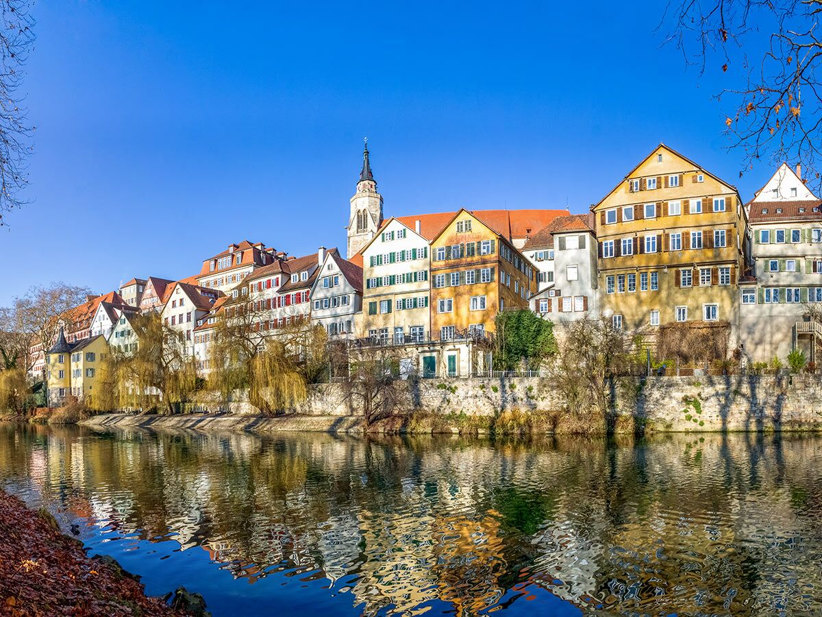 Malerische Neckarfront in Tübingen mit bunten Häusern, Stiftskirche und ihren Spiegelungen im Neckar unter blauem Himmel.