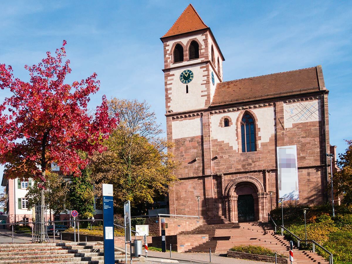 Historische Kirche aus rotem Sandstein mit Uhrturm und Treppenaufgang, umgeben von Herbstbäumen mit rotem Laub unter blauem Himmel.