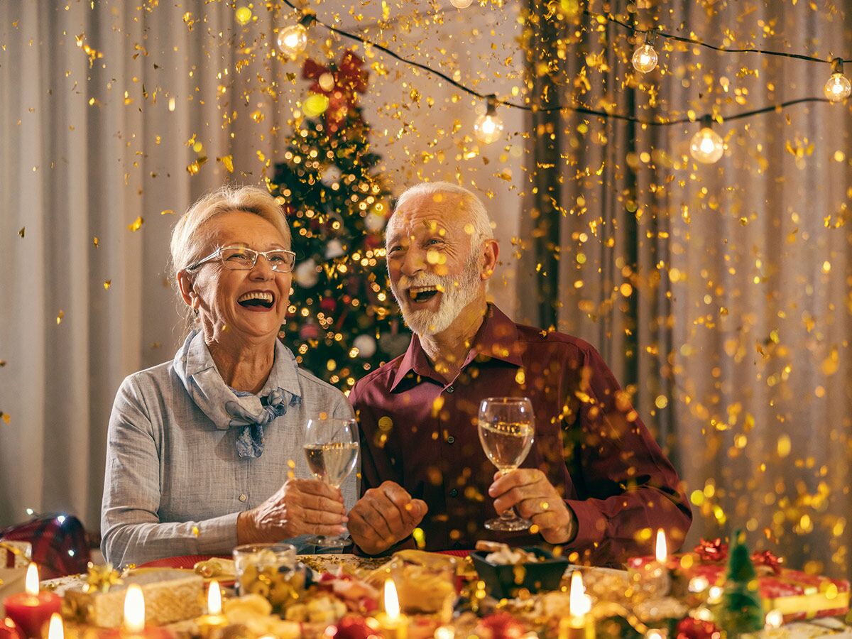Glückliches Senioren-Paar feiert lachend mit Weingläsern an einem festlich dekorierten Tisch unter fallendem Konfetti und Weihnachtsbaum.