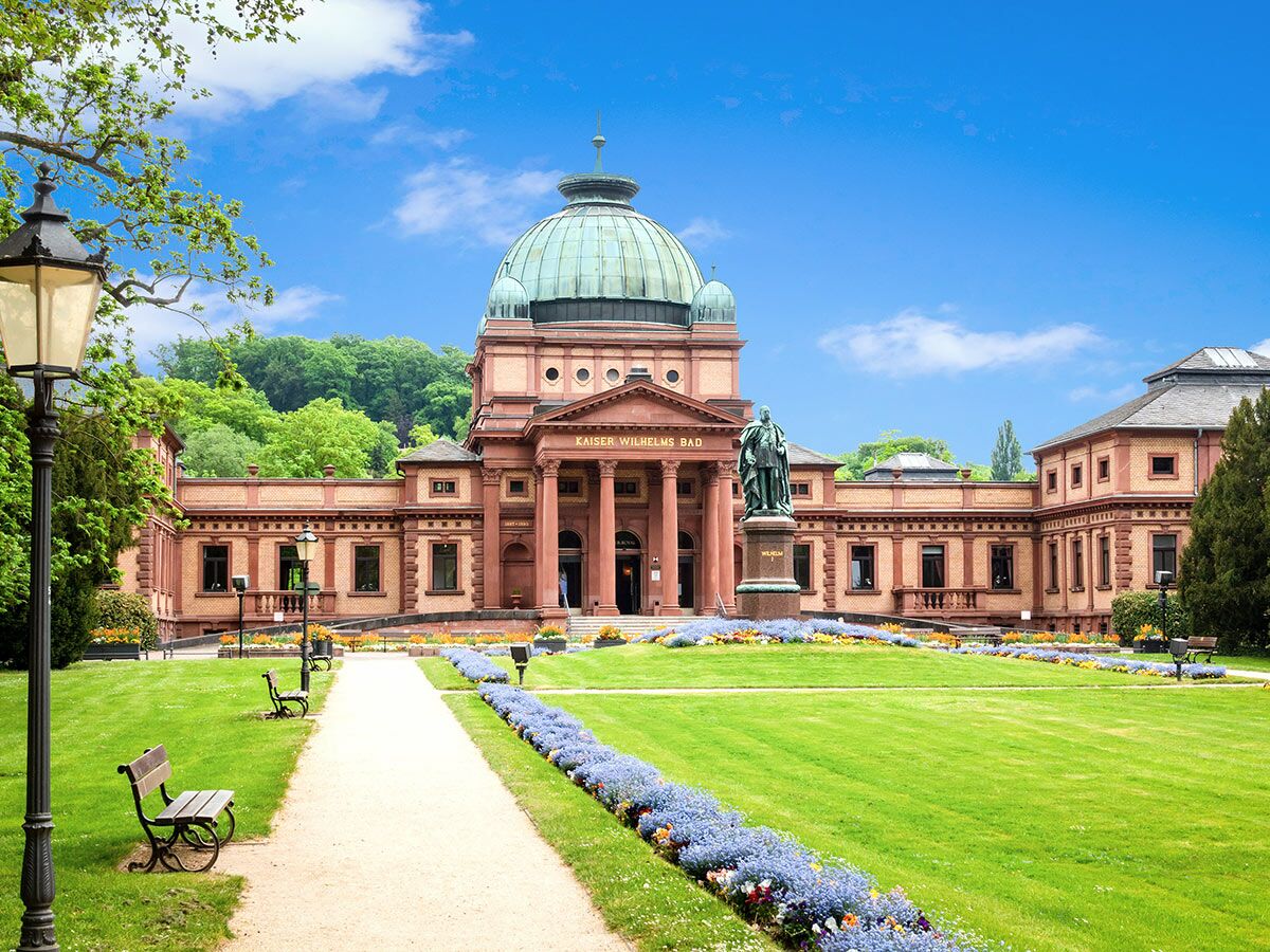 Historisches Kaiser-Wilhelms Bad in Baden-Baden: Prachtvolles Gebäude mit Kuppel, Statue, Park, Wegen, Bänken unter blauem Himmel.