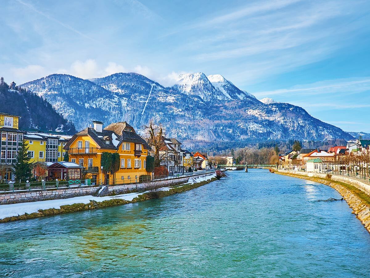 Idyllische Winteransicht von Bad Ischl mit dem Traunfluss, schneebedeckten Bergen und historischen Häusern entlang des Ufers bei Sonnenschein.