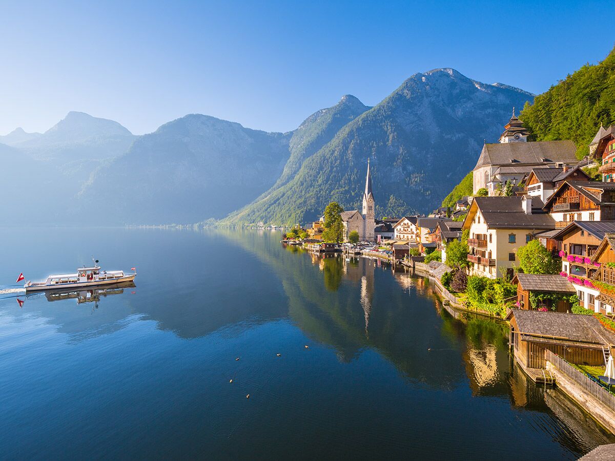 Malerisches Hallstatt Dorf am See in Österreich mit Kirche, traditionellen Häusern und Bergen im Sonnenlicht, ein Boot fährt vorbei.