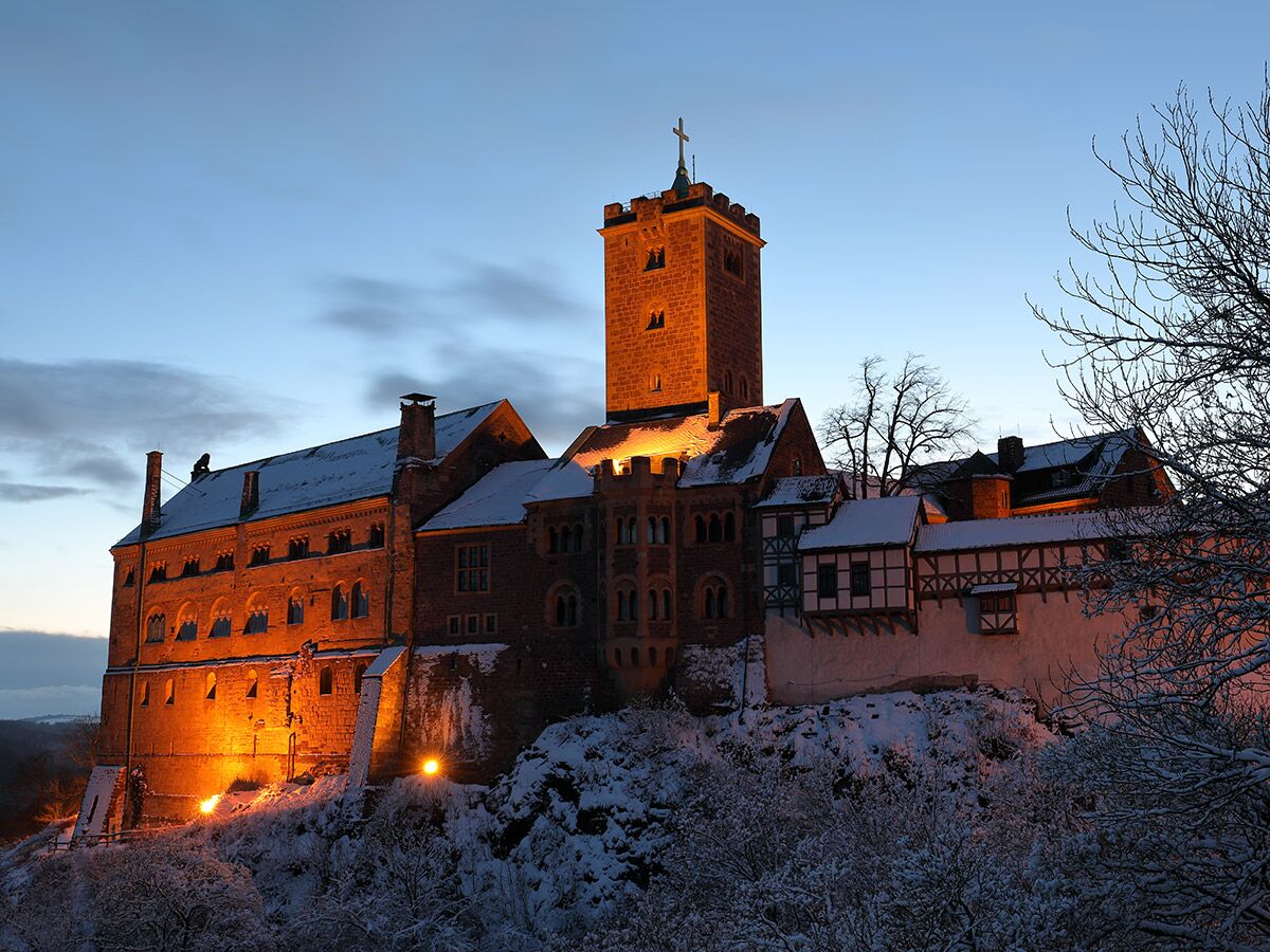 Historische Burg auf verschneitem Hügel in der Dämmerung, warm beleuchtet, blauer Himmel und kahle Bäume.