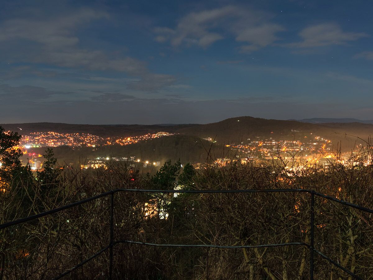 Panoramablick auf eine beleuchtete Stadt in hügeliger Landschaft bei Nacht unter einem sternenklaren Himmel.