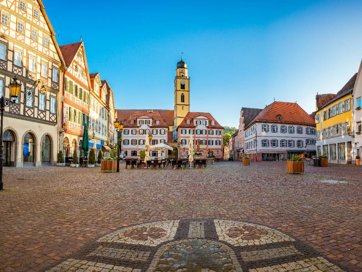 Malerischer deutscher Marktplatz mit Fachwerkhäusern, Uhrturm und leeren Straßencafés am sonnigen Morgen.