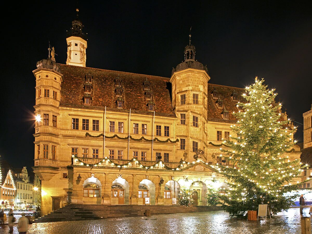 Historisches Rathaus im Schein der Weihnachtsbeleuchtung und eines großen, geschmückten Weihnachtsbaums auf einem Platz bei Nacht.