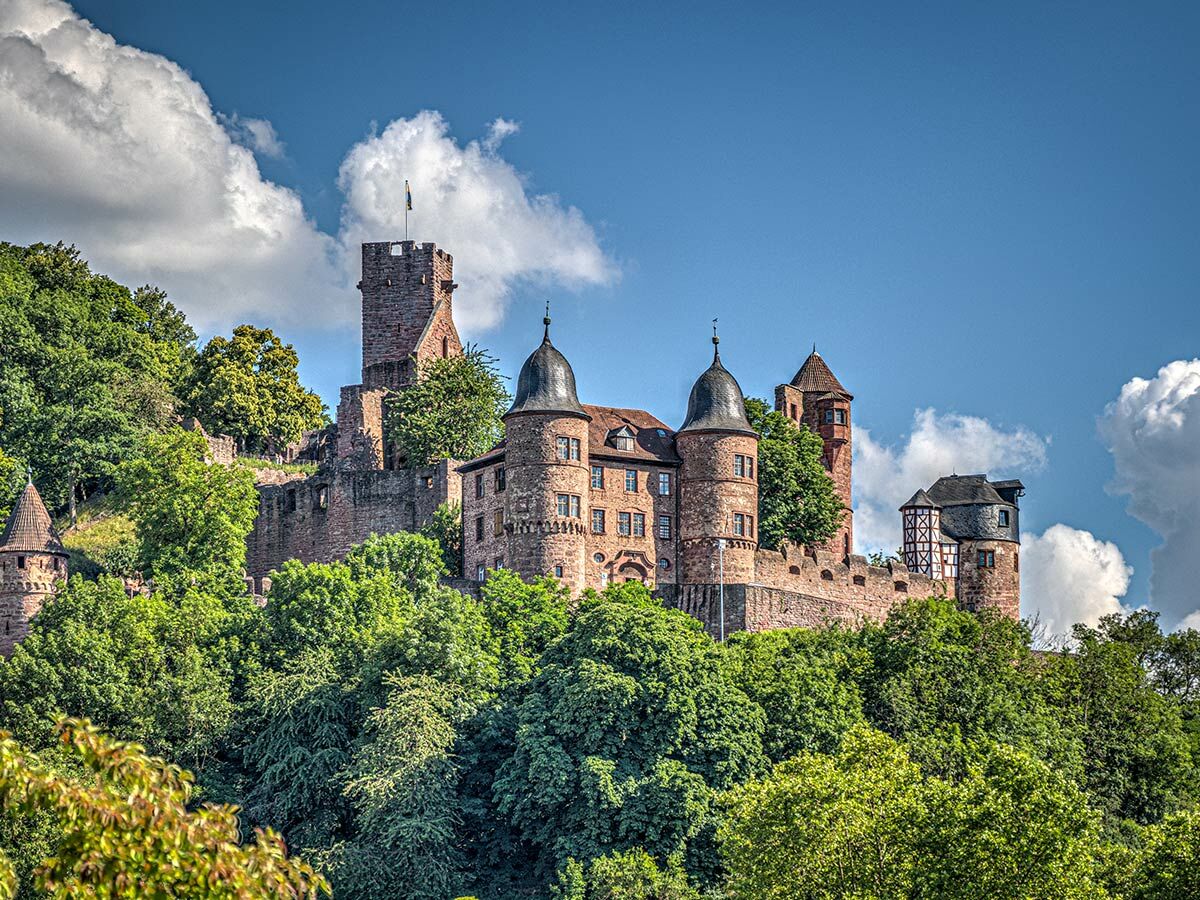 Majestätische deutsche Burg mit Türmen und Mauern auf einem grünen, bewaldeten Hügel unter blauem Himmel mit Wolken.