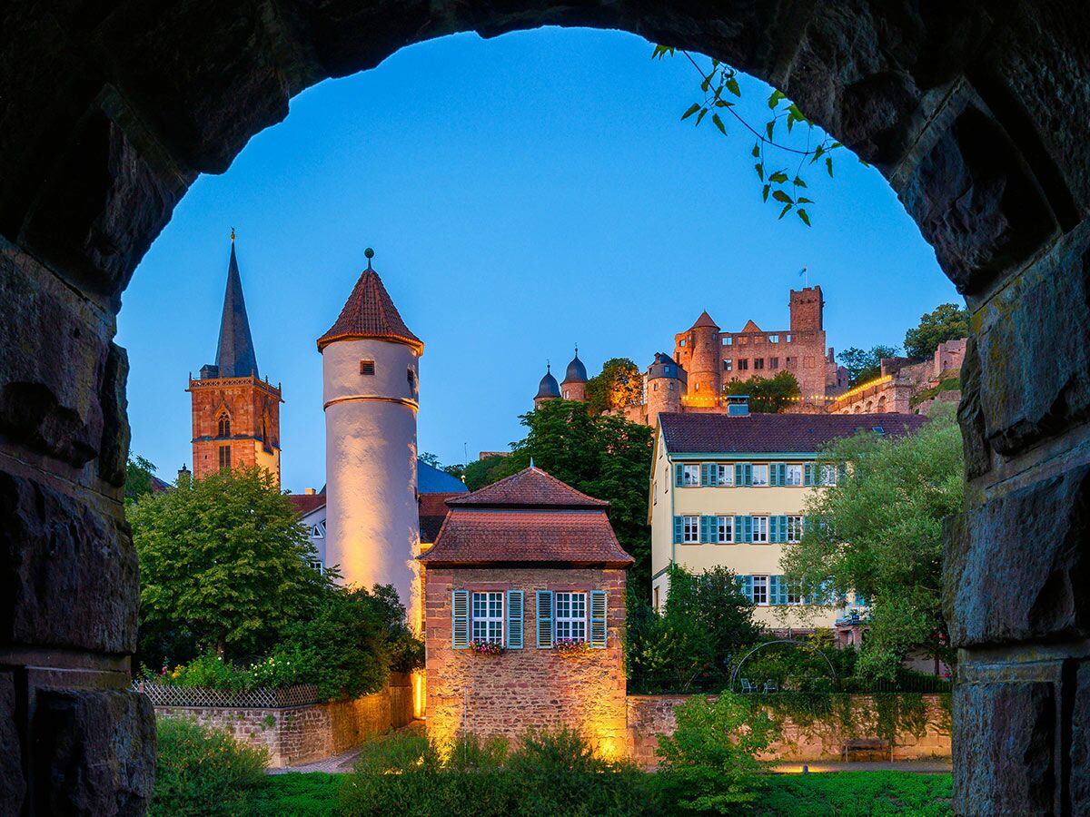 Blick durch Steinbogen auf Gengenbach mit beleuchteter Kinzigtorturm, Abteikirche und Klosteranlage auf dem Berg in der Abenddämmerung.