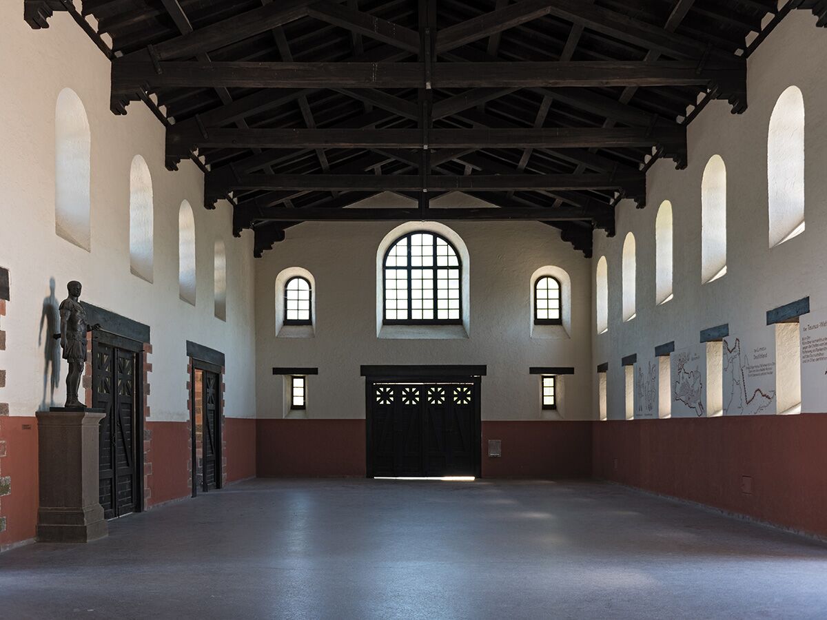 Geräumiger historischer Saal mit römischer Soldatenstatue, hohen Bogenfenstern, dunkler Holzbalkendecke und hellen Wänden.